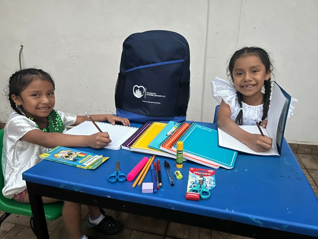 En imagen dos niñas de comunidades de Oaxaca sonrién mientras escriben y al fondo una mochila de útiles con el logotipo de la fundación Arnoldo Janssen